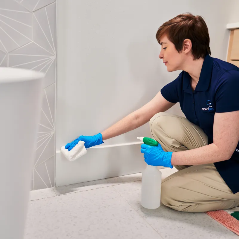 Professional cleaner wearing gloves wipes wall near floor with spray bottle in modern bathroom
