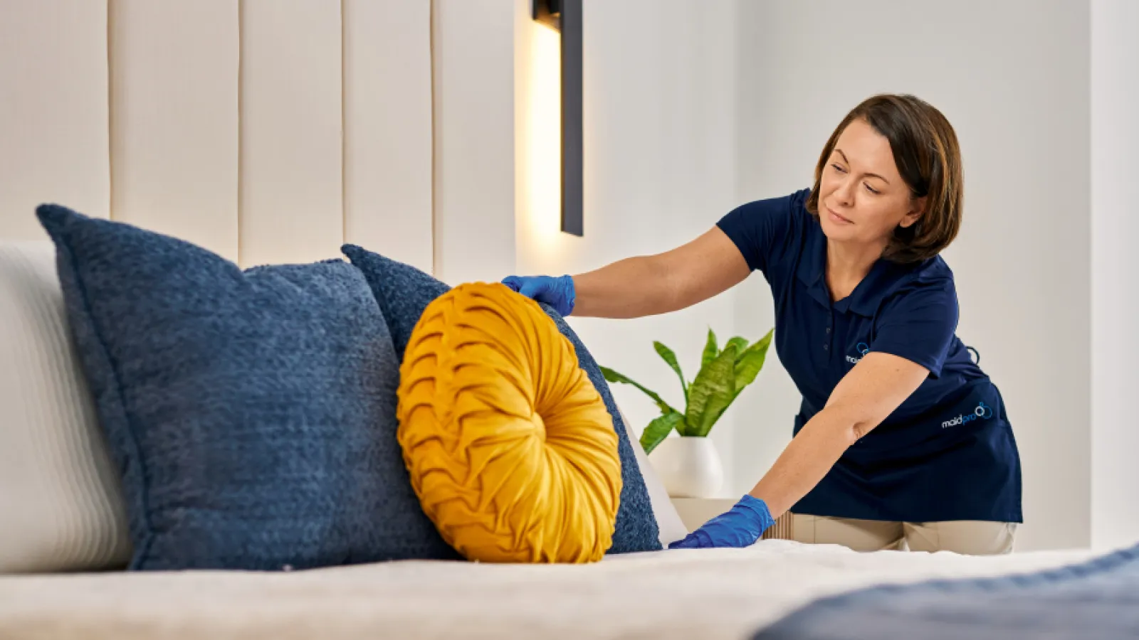 Professional cleaner arranging mustard yellow and navy blue pillows on a bed in a modern bedroom.