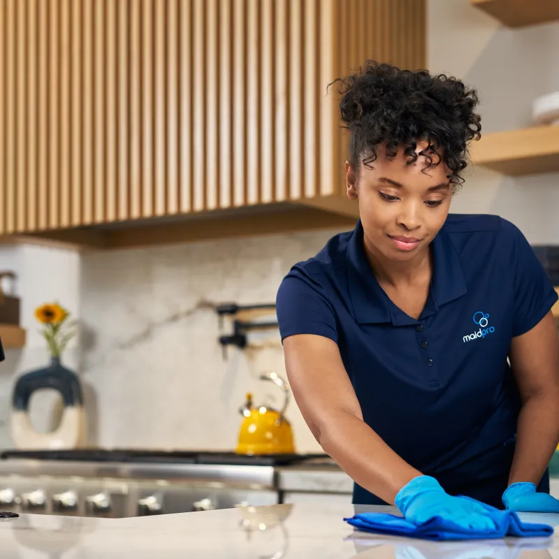 Woman in blue uniform cleaning kitchen countertop with cloth and spray bottle wearing protective gloves.